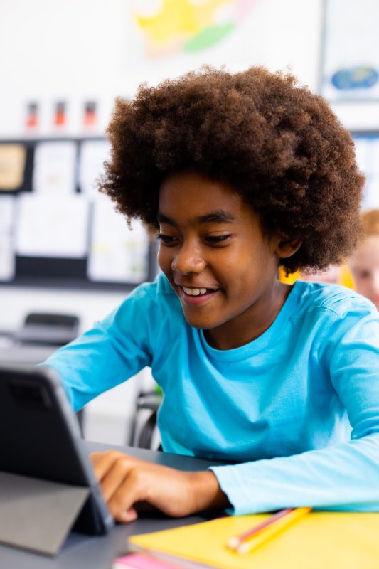 Happy african american schoolboy sitting at desk and using tablet in school classroom. School, education and learning concept. Happy african american schoolboy sitting at desk and using tablet in school classroom. School, education and learning concept.